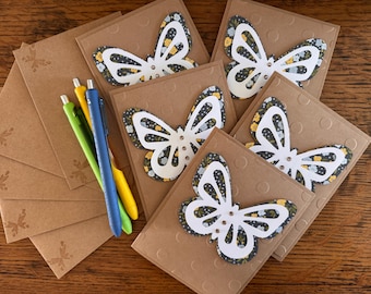 May include: Set of six brown cardstock cards with white butterfly cutouts. Each card has a floral pattern in shades of yellow, white, and black. The cards are arranged in a fan shape with three pens on top.