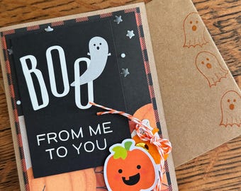 May include: A Halloween card with a black background and the words "Boo! From me to you". The card has a ghost and a pumpkin with a happy face on it. The card is tied with a red and white string.