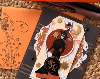 May include: A handmade greeting card featuring a witch illustration in a black dress and hat, with a circular 'BOO' emblem and an 'ALL DRESSED UP' banner. The card includes layered paper elements, gold star accents, and a matching orange envelope with a broom and boot stamp.