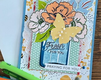 May include: A handmade greeting card with a floral design and a yellow butterfly. The card features the text "Fresh for You" and "Praying for You". The card is decorated with a variety of colors, including orange, pink, and blue. Pens in green, orange, and blue are in the foreground.