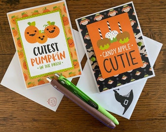 May include: Two Halloween-themed greeting cards. The first card features a pumpkin design with the text "Cutest Pumpkin in the Patch." The second card features a candy apple design with the text "Candy Apple Cutie."