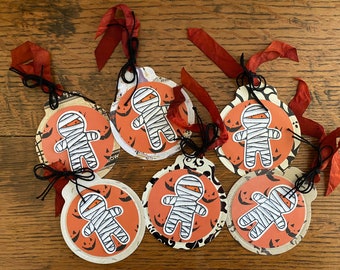 May include: Six Halloween-themed gift tags featuring a mummy gingerbread man design. Each tag is decorated with orange, black, and white colors and has a black ribbon attached.