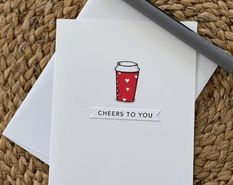 May include: A white greeting card with a red coffee cup illustration and white hearts. The card reads "Cheers to You" and is accompanied by a white envelope and a gray pen. The card is on a woven, tan surface.