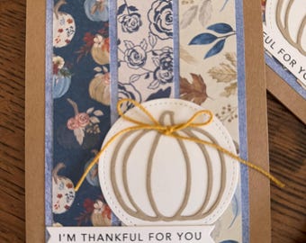 May include: Handmade fall greeting cards with a pumpkin design. The cards feature a white pumpkin with a gold string, set against patterned paper in shades of blue, brown, and cream. The text "I'm thankful for you" is printed on a banner.