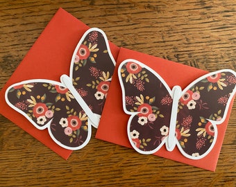 May include: Two butterfly-shaped greeting cards with floral patterns, resting on orange envelopes. The butterflies have a dark background with red, pink, and yellow flowers. The envelopes are a solid orange color, and the background is a wooden surface.