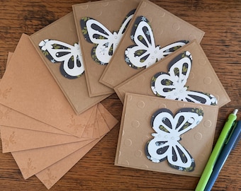May include: A collection of handmade greeting cards and envelopes. Each card features a white butterfly design with black and floral accents, set against a textured brown background. Envelopes are a matching brown color. Two pens, green and blue, are also visible.