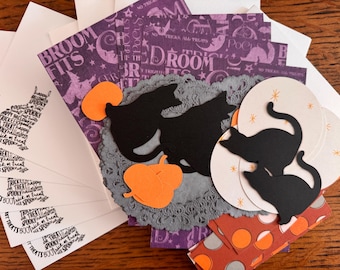 May include: A collection of Halloween-themed paper craft supplies including purple patterned cardstock with text, black cat silhouettes, orange pumpkin cutouts, white circular tags with star accents, and white cards featuring cat-shaped text.