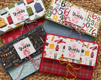 May include: Four colorful school-themed gift card holders. Each features a patterned design with school supplies, backpacks, and numbers. A white square with the word "thanks" and a schoolhouse image is tied with twine.