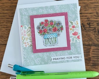 May include: A handmade greeting card with a floral illustration and the text "Praying for You." The card features a bouquet of red roses and white daisies in a blue vase. Two pens, one green and one blue, are resting on the card.
