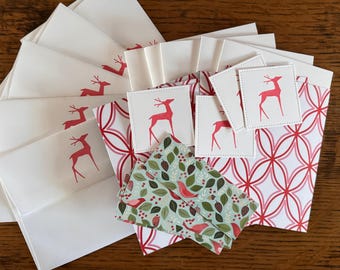 May include: A collection of stationery items, including white envelopes with red deer silhouettes, square cards with deer designs, and patterned paper with red and white geometric shapes and a bird and leaf pattern. The items are arranged on a wooden surface.