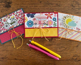 May include: Three handmade greeting cards with decorative paper and embellishments. One card features a bicycle with flowers, another a camper, and the third a sun and cloud. Two pens, one yellow and one pink, are also visible.
