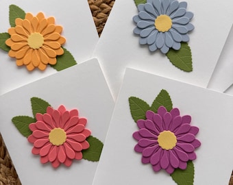 May include: Four white greeting cards, each adorned with a colorful, layered paper flower and green leaves. The flowers are in shades of orange, blue, pink, and purple, with yellow centers. The cards are arranged on a textured surface.
