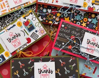 May include: A collection of handmade thank you cards with school-themed patterns. Each card features a white square with the word "thanks" and a schoolhouse illustration. The cards have various designs, including pencils, scissors, and school buses.