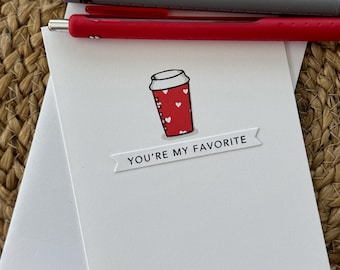 May include: A white greeting card with a red coffee cup design and the text "YOU'RE MY FAVORITE." The coffee cup has white hearts. Two pens, one gray and one red, are on top of the card. The card is on a woven surface.