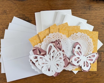 May include: A collection of handmade greeting cards and envelopes. The cards feature white paper butterflies, doilies, and geometric patterned paper in shades of pink and yellow. White envelopes are stacked to the left.