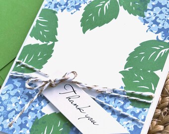 May include: A rectangular greeting card featuring a blue hydrangea floral pattern with green leaves on a white background. A white tag with the text Thank you is attached with a metallic gold and white twine bow, paired with a green envelope.