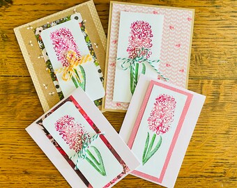May include: Four handmade greeting cards with pink hyacinth flowers and green stems. Each card has a different patterned background and is tied with twine.