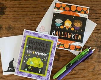 May include: Two Halloween greeting cards. The first card has a purple background with bats and clouds. The text on the card reads "Bubble Bubble Toil & Trouble". The second card has a black background with a row of three children dressed as a vampire, a witch, and a werewolf. The text on the card reads "Halloween".