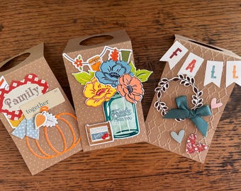 May include: Three brown paper gift bags with die-cut handles. The left bag features a pumpkin, a red patterned shape, and the text "Family together". The center bag shows a mason jar with flowers and "Fresh for you". The right bag has a "FALL" banner, a wreath, and a bow.