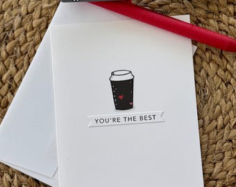 May include: A white greeting card with a black coffee cup illustration and the text "YOU'RE THE BEST". A gray and red pen rest on top of the card and envelope, all on a woven surface.