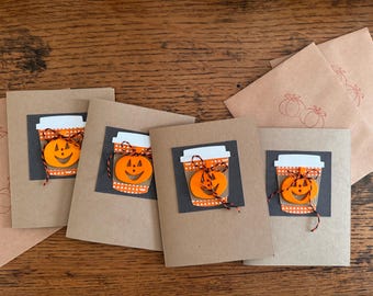 May include: Four brown cards with a black and white pumpkin-shaped coffee cup design. Each card has a different pumpkin face. The cards are on a wooden surface.