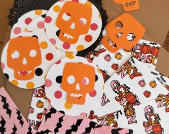 May include: A collection of Halloween-themed craft supplies. Includes orange skull cutouts, polka dot circles with skulls, pink bat-patterned strips, and envelopes with pumpkin and "October 31st" designs. A black lace doily is also present.