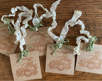 May include: Five brown paper gift tags with a brown ink illustration of a pumpkin and vine. Each tag has a hole punched in the top and is tied with a white satin ribbon and a green twine bow.