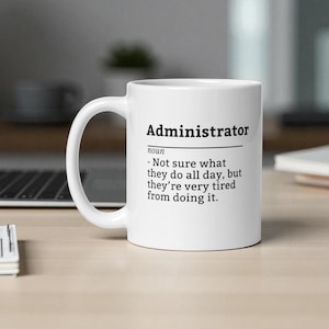 May include: White ceramic mug with the word "Administrator" printed in black, along with a humorous definition. The mug has a curved handle and is set on a light-coloured surface.