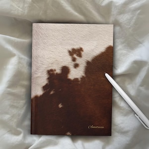 Puede incluir: Un cuaderno de tapa dura con un diseño de piel de vaca en tonos marrones y blancos. El nombre "Anastasia" está impreso en oro en la esquina inferior derecha. Un bolígrafo blanco descansa al lado.