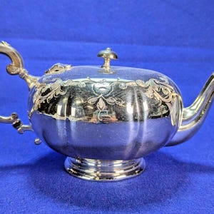 May include: A polished silver teapot featuring a rounded, fluted body with an ornate engraved floral pattern along the upper rim. It includes a curved handle, a slender spout, a circular base, and a small lid with a decorative knob handle on top.