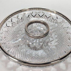 May include: A round clear glass serving dish featuring a complex diamond-cut pattern and a central circular compartment. The dish is finished with a polished silver-tone metal rim and a matching silver-tone metal ring around the center section.