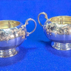May include: A silver-toned sugar bowl with two handles and a matching creamer pitcher with a single handle and spout. Both vessels feature a rounded, fluted body with an ornate, engraved floral scroll pattern around the upper rim and a pedestal base.