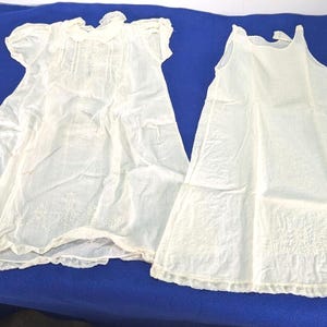 May include: Two off-white sleeveless and short-sleeved dresses made of lightweight fabric. The left dress features a rounded collar, vertical pleats on the bodice, and delicate embroidery near the hem. The right dress has a simple scoop neckline.