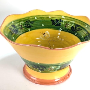 Puede incluir: Un cuenco decorativo de cerámica amarilla con borde festoneado. El cuenco presenta una banda verde con diseños florales y una base naranja claro. El interior es amarillo con una mariposa pintada a mano.