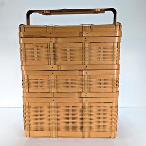 May include: A rectangular, light brown woven bamboo basket featuring a tiered, stacked design with three distinct sections. The basket includes a sturdy, arched handle at the top and is constructed with vertical and horizontal woven strips.