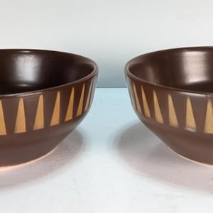 Könnte beinhalten: Zwei braune Keramikschalen mit matter Oberfläche. Jede Schale hat ein Band aus sich wiederholenden goldfarbenen Dreiecken an der Außenseite. Die Schalen haben ein schlichtes, modernes Design.