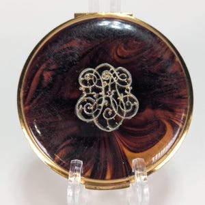May include: A round compact mirror with a gold-toned metal frame and a dark brown and black marbled resin lid. Centered on the lid is an intricate, silver-toned metal filigree ornament featuring a swirling, decorative pattern of interconnected loops.