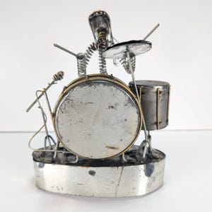 May include: A metal sculpture of a drummer playing a drum kit. The figure is constructed from wire and springs, positioned behind a large circular bass drum and a smaller cylindrical tom drum. A cymbal and a microphone stand are also included.