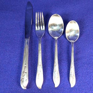 Puede incluir: Un juego de cuatro cubiertos plateados: un cuchillo, un tenedor y dos cucharas. Los mangos tienen un diseño floral. Los cubiertos están colocados sobre una superficie azul.