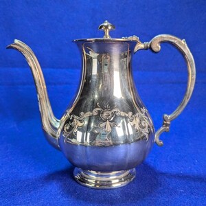 May include: A silver-toned metal teapot featuring a curved spout, a hinged lid with a small finial, and an ornate handle. The body of the teapot is decorated with an engraved floral pattern and has a rounded base that tapers toward the top.
