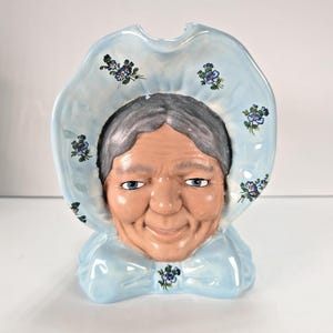 May include: A ceramic head vase featuring a sculpted face with gray hair and blue eyes, framed by a light blue ruffled bonnet decorated with small blue floral patterns. The piece has a glossy finish and a wide, scalloped opening at the top.