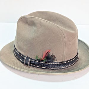 May include: A taupe fedora hat featuring a black textured leather band with light-colored stitching. The hat is accented with a small decorative cluster of feathers in shades of red, green, and black tucked into the side of the hat band.