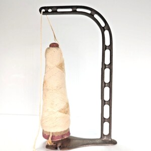 May include: A dark metal thread stand with a curved arm featuring rectangular cutouts. A tall, conical spool of off-white thread sits on the base, with a single strand of thread extending from the top of the spool through the arm's eyelet.