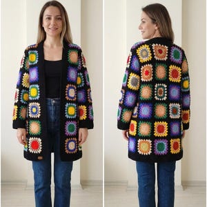May include: A long, open-front crochet cardigan in a patchwork design. The cardigan features a black base with colorful squares in shades of yellow, blue, purple, green, and orange. The sleeves and edges are trimmed in black.
