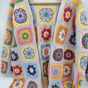 Può includere: Un cardigan all'uncinetto aperto e colorato. Il capo presenta un design patchwork di motivi quadrati in vari colori, tra cui rosa, giallo, blu e verde, con un bordo grigio neutro. Il cardigan è esposto su una gruccia di legno.