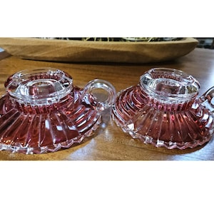 May include: Two matching pink glass candle holders featuring a circular base with a radial ribbed pattern and a small side handle. The center of each holder has a clear, smooth glass rim designed to securely hold a standard taper candle.