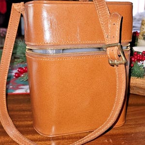 May include: A rectangular, tan leather handbag with a zipper closure and a long, adjustable shoulder strap. The bag has a structured design with a buckle detail on the strap. The bag is approximately 10 inches tall.
