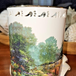 May include: White ceramic mug with a landscape scene featuring a fence, trees, and colorful flowers. The rim has a gold trim and decorative cutouts. A white handle is attached to the side.