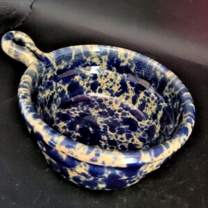 May include: A small, round ceramic bowl with a single handle, featuring a speckled pattern of deep navy blue and creamy beige. The interior and exterior surfaces are glazed with a glossy finish, showing a mottled, sponge-painted appearance.