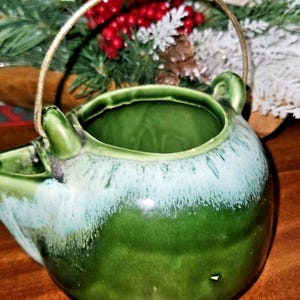 May include: A green ceramic teapot with a gold handle. The teapot has a rounded shape with a spout and a handle. The top of the teapot has a light blue glaze, and the rest of the teapot is a dark green color.